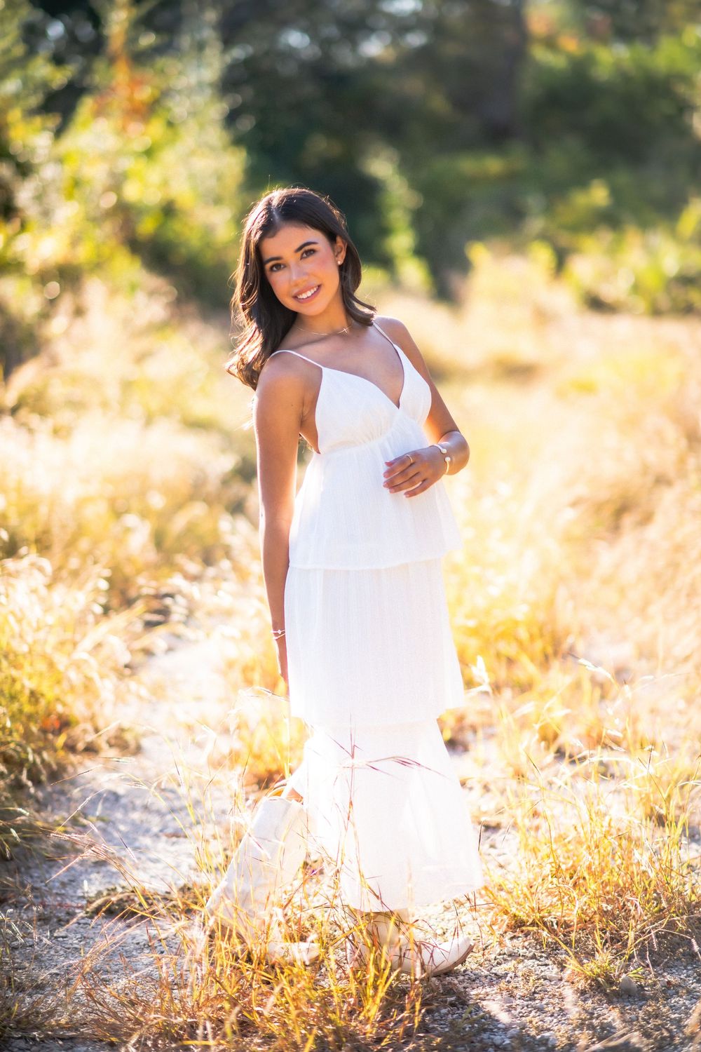 Smiling pregnant woman in white dress standing in sunlit field with golden grass and green trees in background.