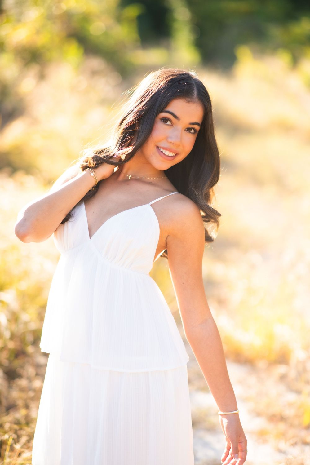 Young woman in white sundress poses outdoors in golden sunlight, smiling over shoulder with long dark hair.