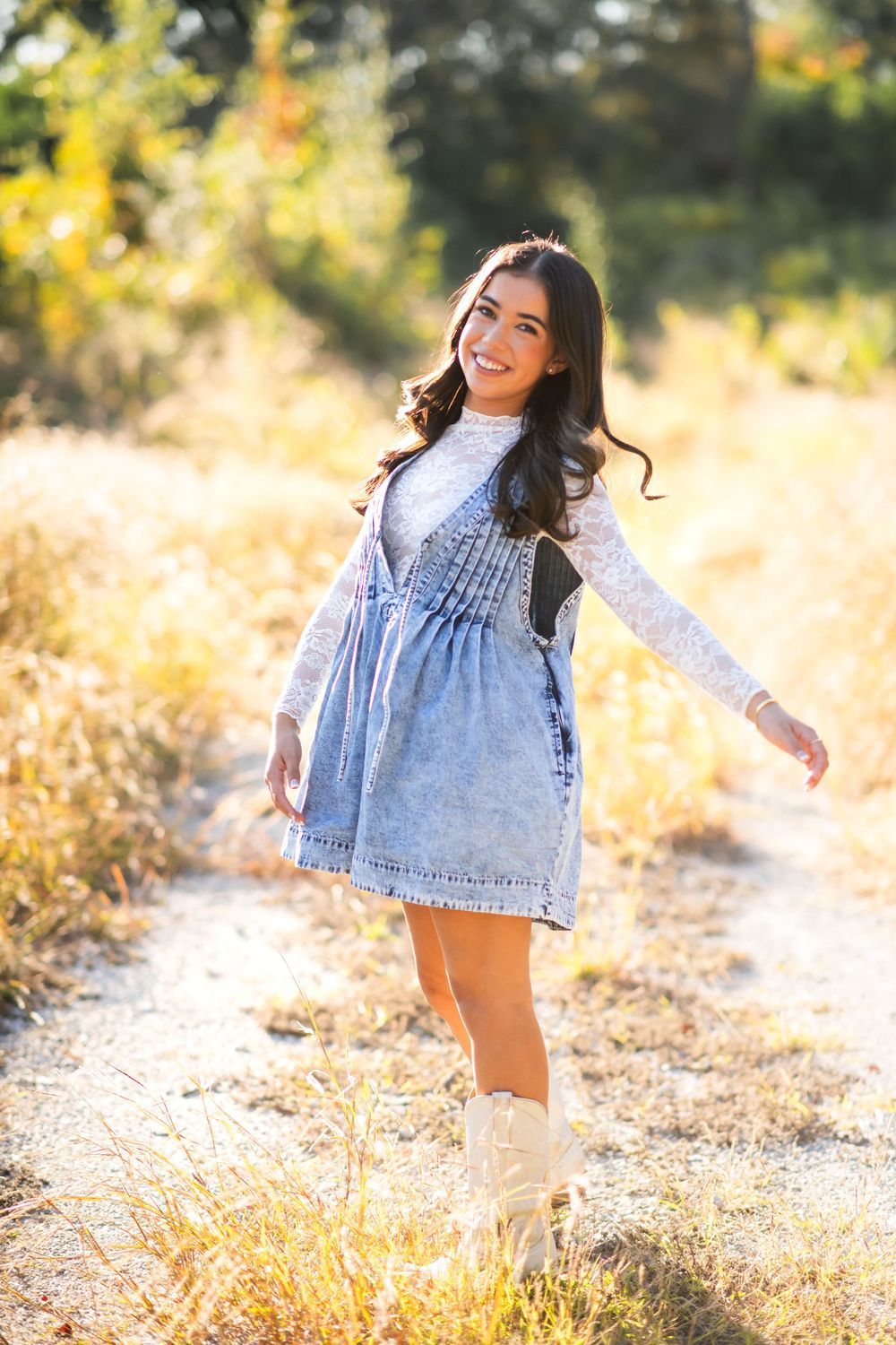 Smiling young woman in denim dress, lace top, and white boots posing on a sunny outdoor path surrounded by golden foliage.