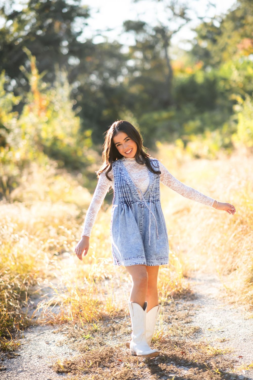 Young woman in denim dress, lace top, and white cowboy boots smiling on a sunny rural path.