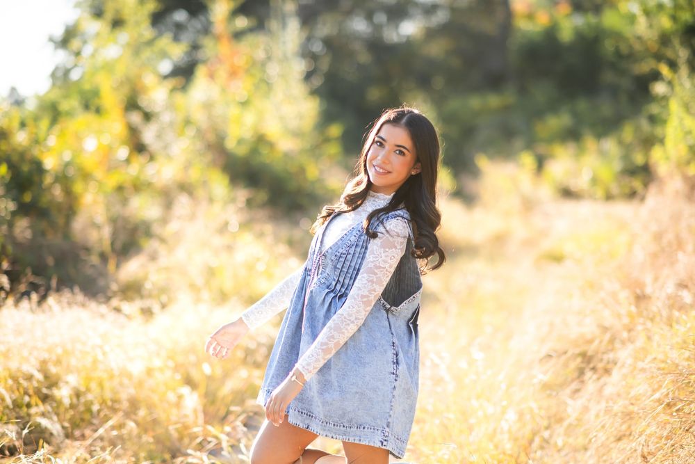 Young woman in denim dress smiling in golden field surrounded by autumn foliage on sunny day.