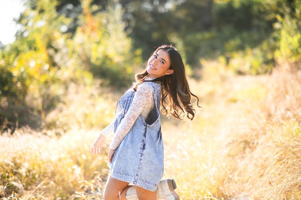Young woman in denim dress smiling over shoulder in sunny golden field with lush green trees backdrop.