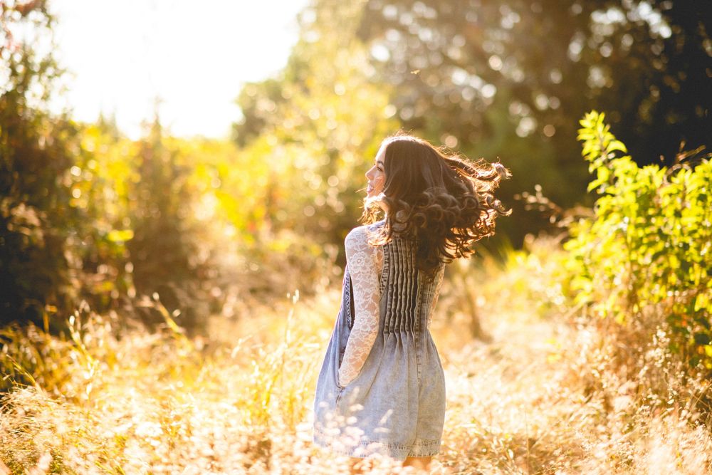 A woman in a lace dress stands in a sunlit meadow, her hair flowing as golden light filters through autumn trees.