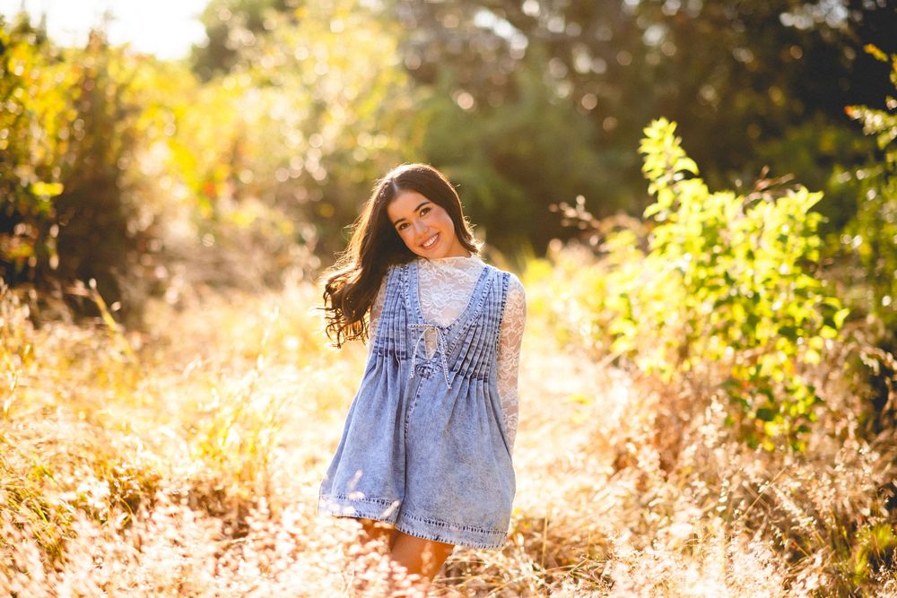 Young woman in blue lace dress smiling in golden sunlit field surrounded by tall grass and green foliage.
