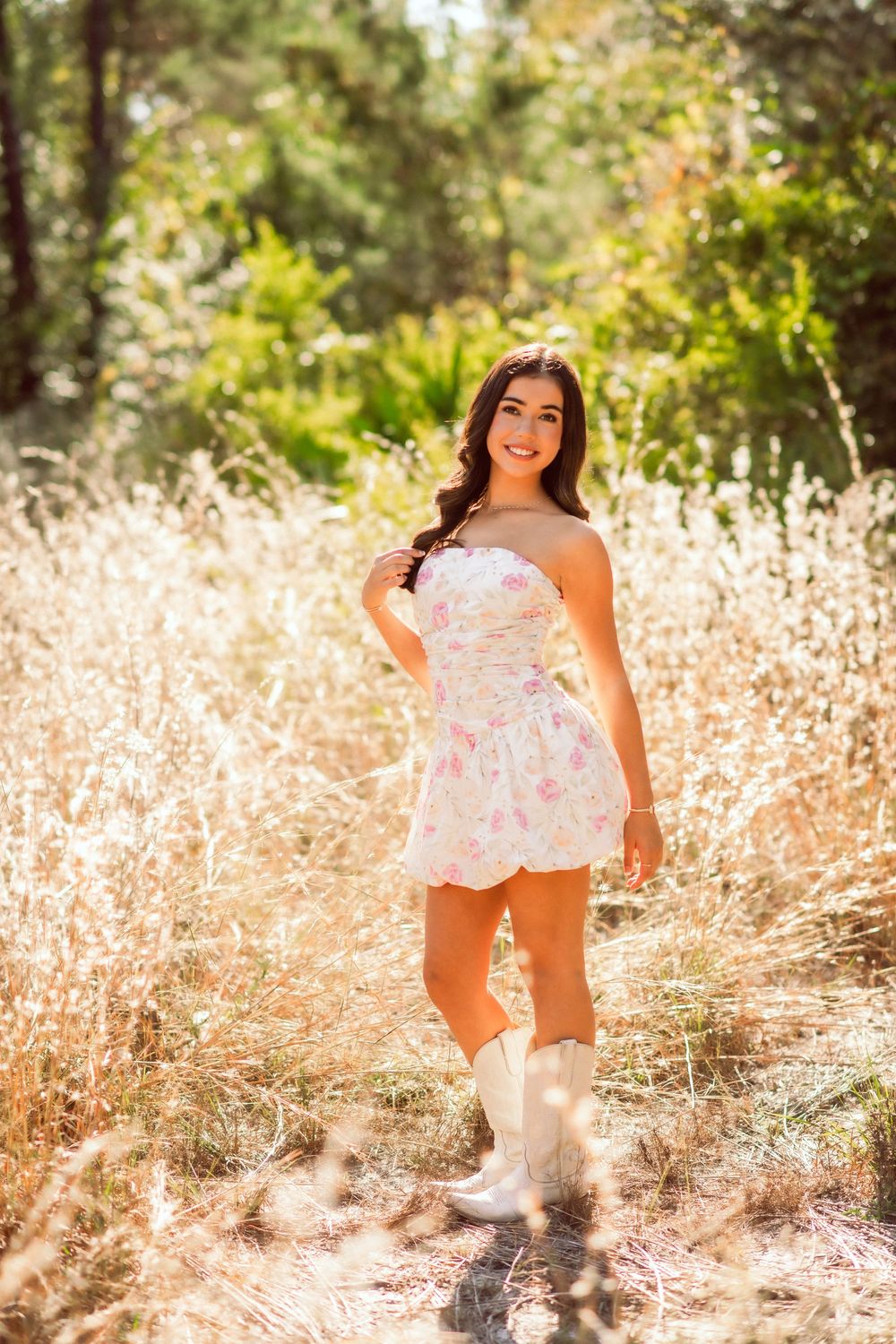 Young woman in floral strapless mini dress and white cowboy boots smiling in a sunlit golden field.