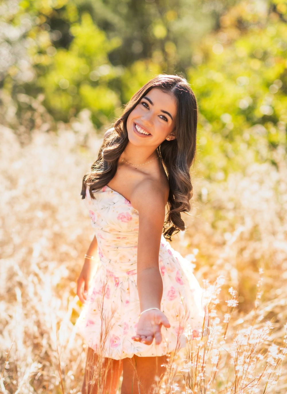 Young woman in floral dress smiling in golden sunlit field, twirling gracefully among tall grasses.