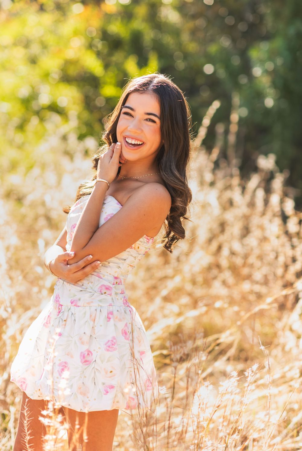 Young woman in floral dress smiling joyfully in a sunlit golden field, posing for a senior portrait photo.