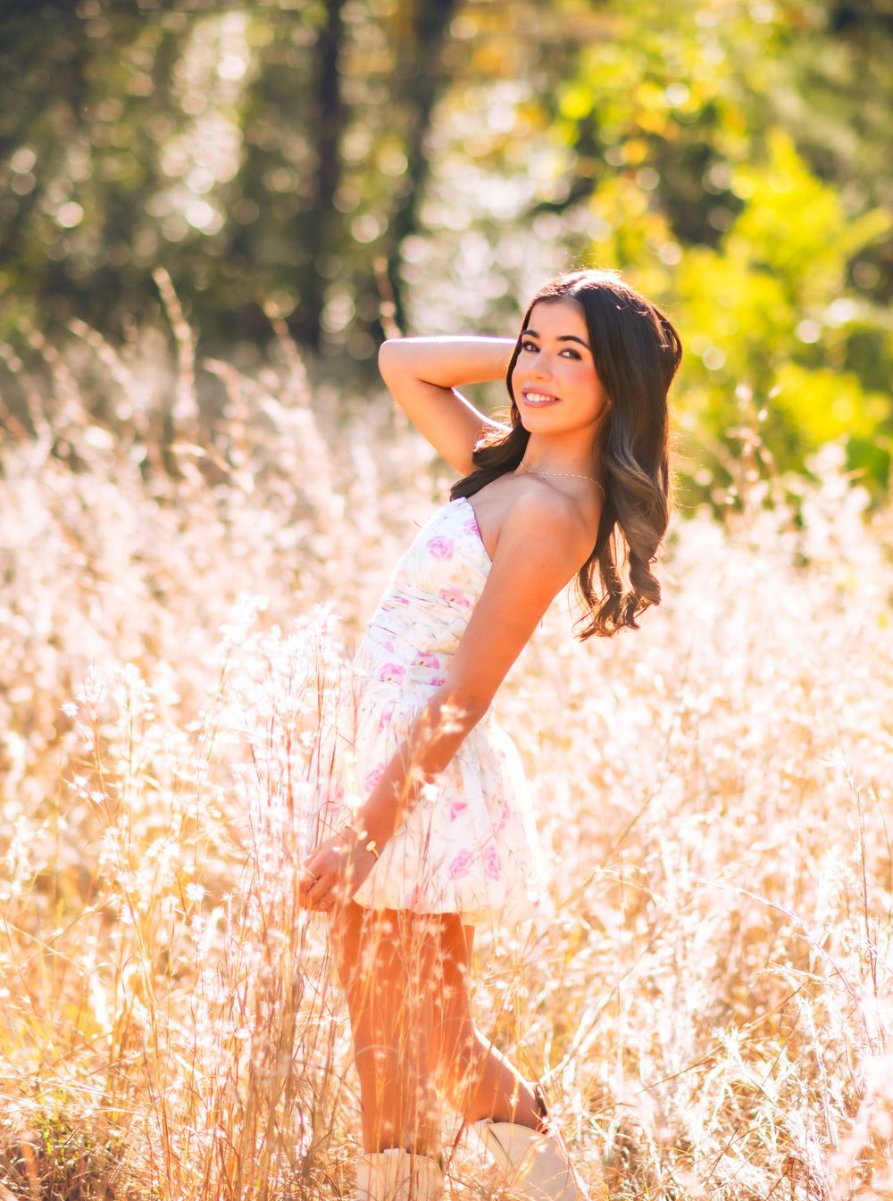 Young woman in floral dress standing in golden sunlit field, smiling over shoulder, with trees in background.