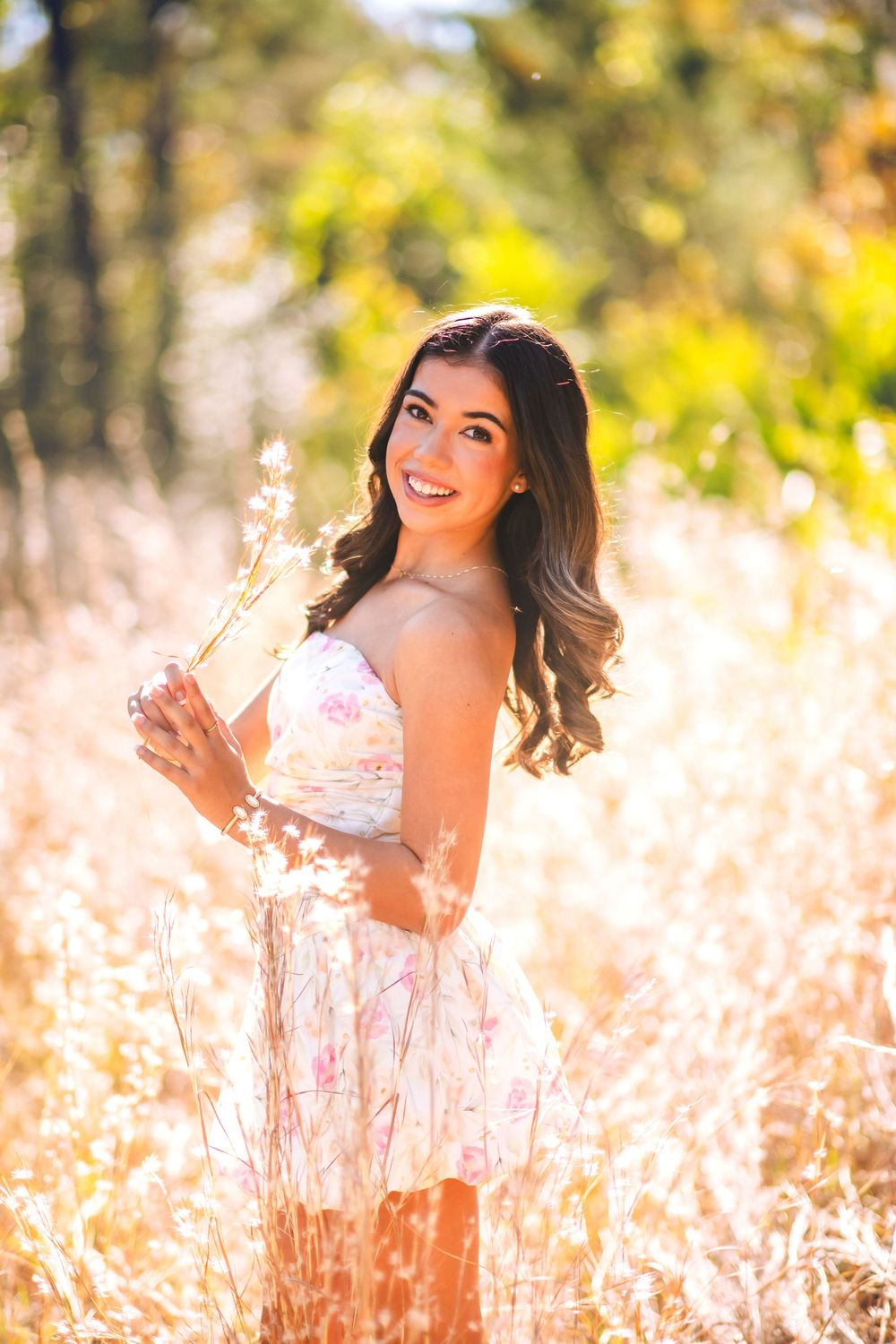 Young woman in floral dress smiling in golden sunlit field holding wildgrass, autumn trees in background.