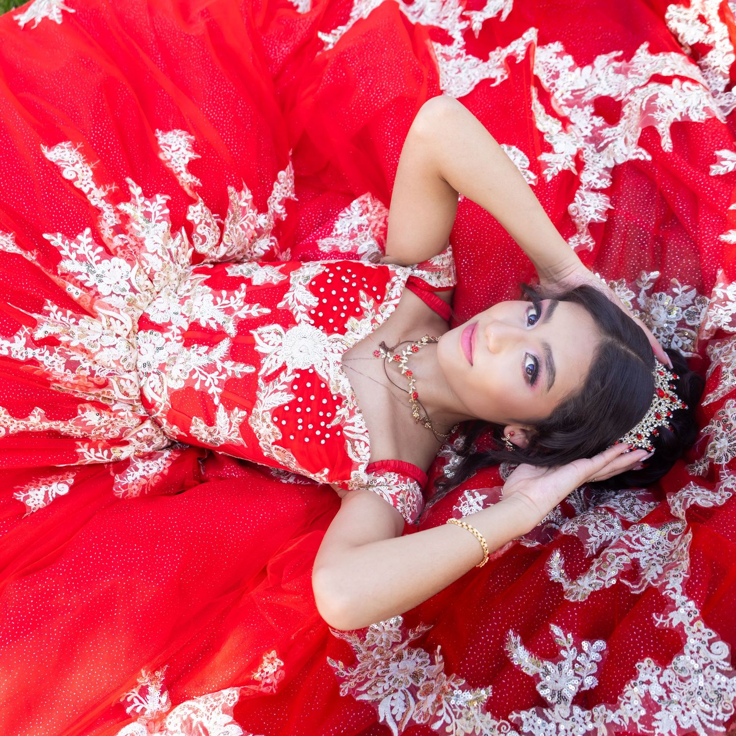 Young woman in elegant red and white lace ball gown lying gracefully, wearing jewelry and a tiara.