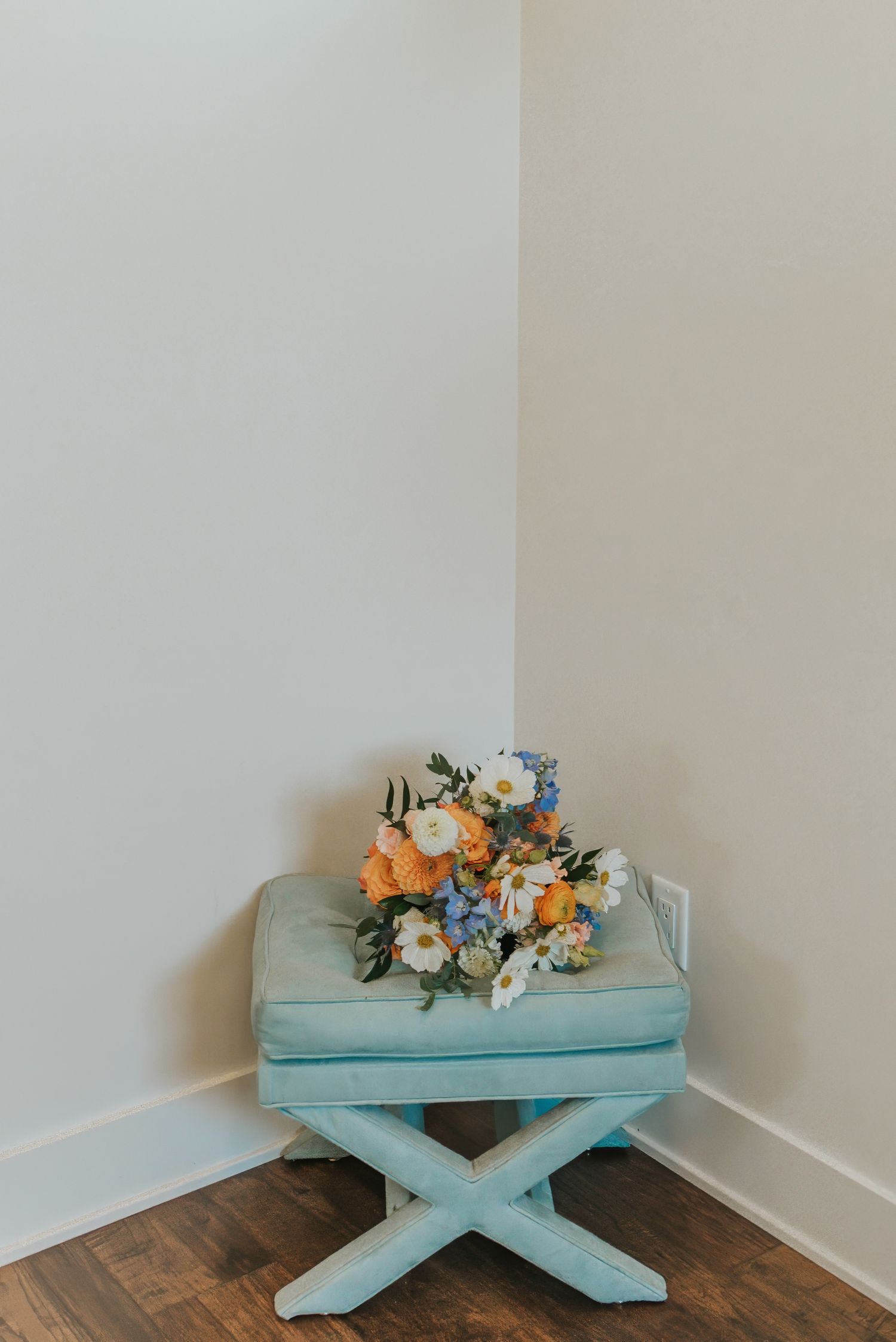 A colorful wedding bouquet with orange, white, and blue flowers resting on a mint green X-base ottoman in a corner.