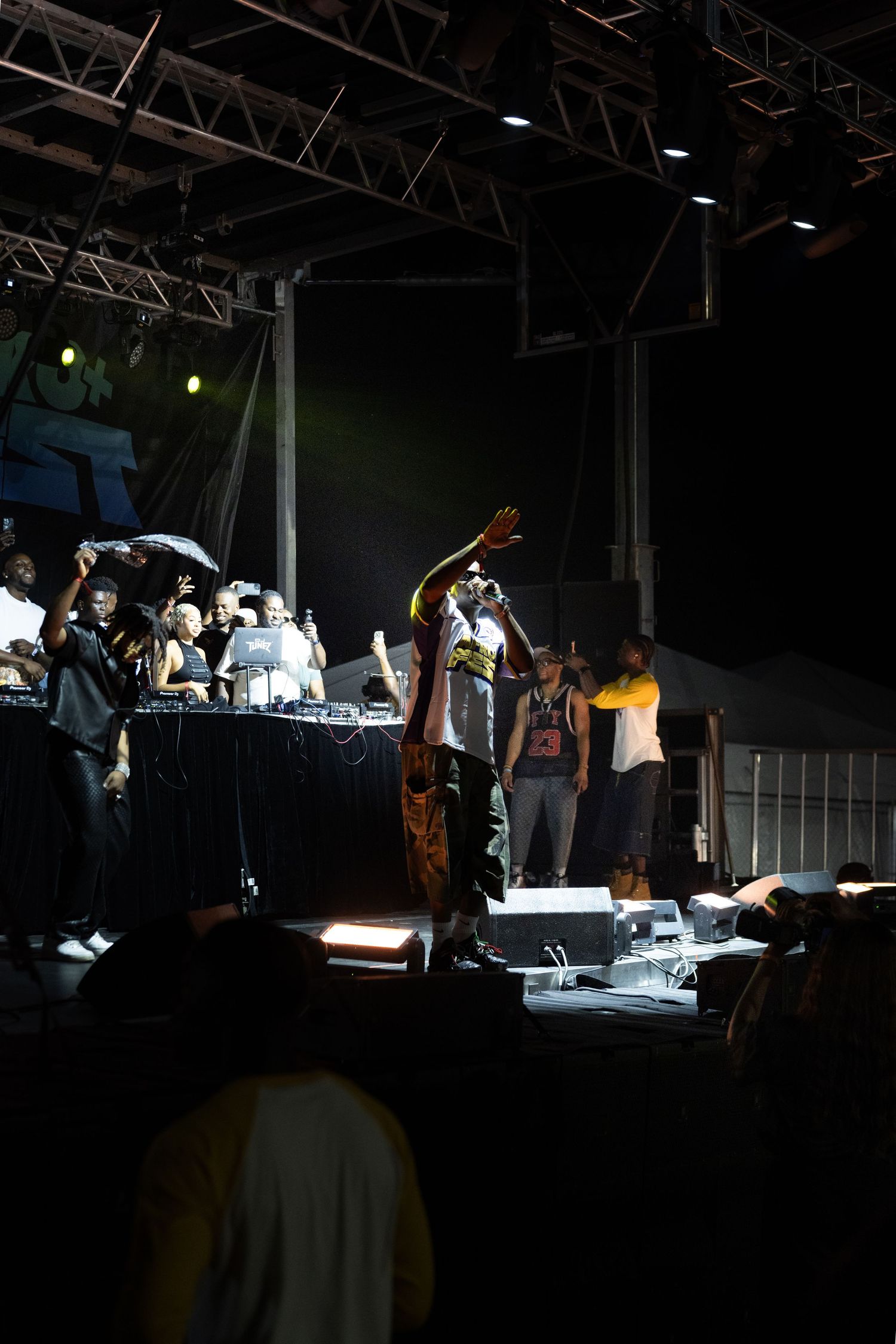 Performers on a dark outdoor concert stage with hands raised, crowd and DJ setup visible in background at night.