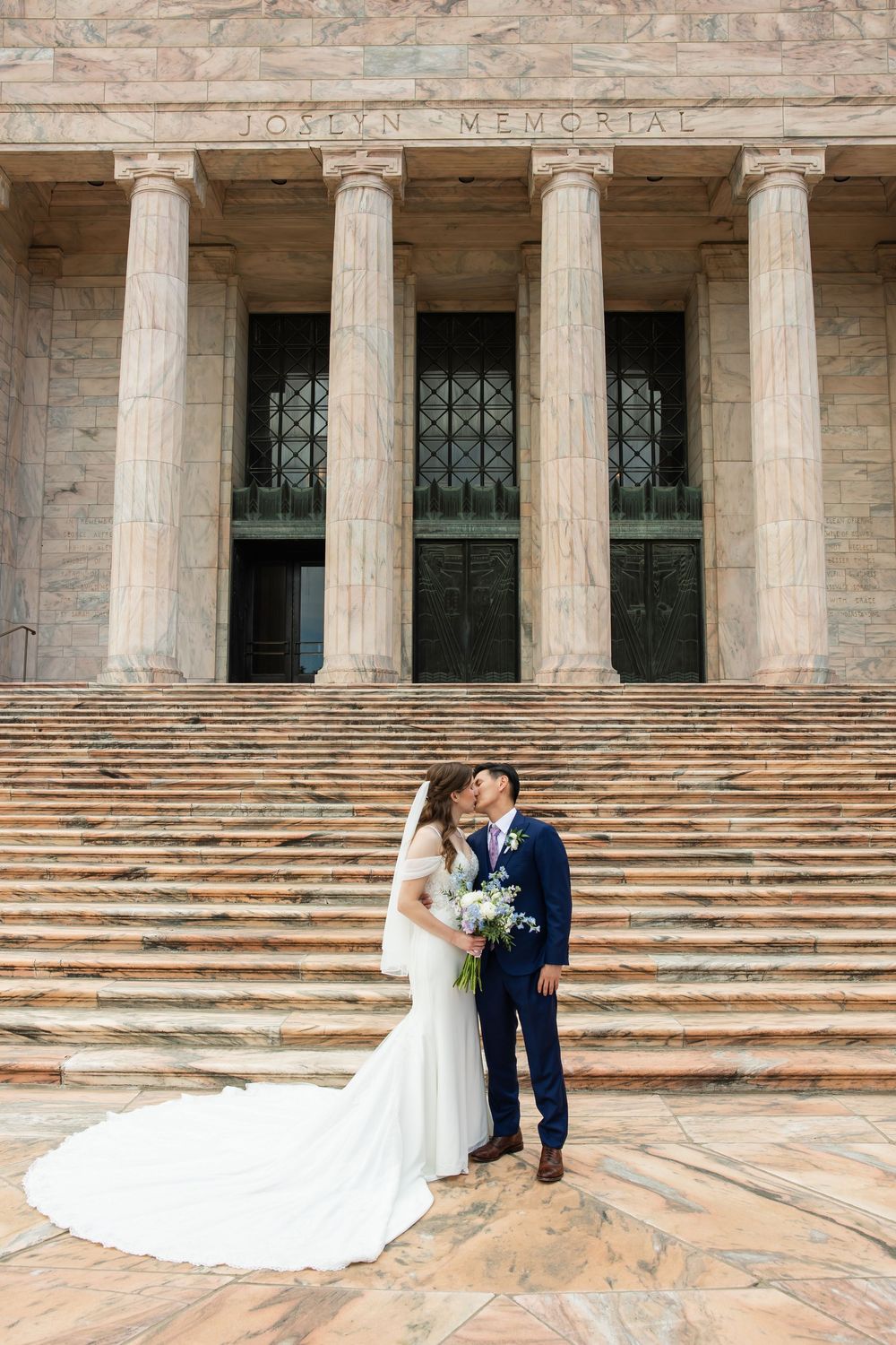 Bride and groom share a kiss on the steps of Joslyn Memorial, bride in white gown holding bouquet.