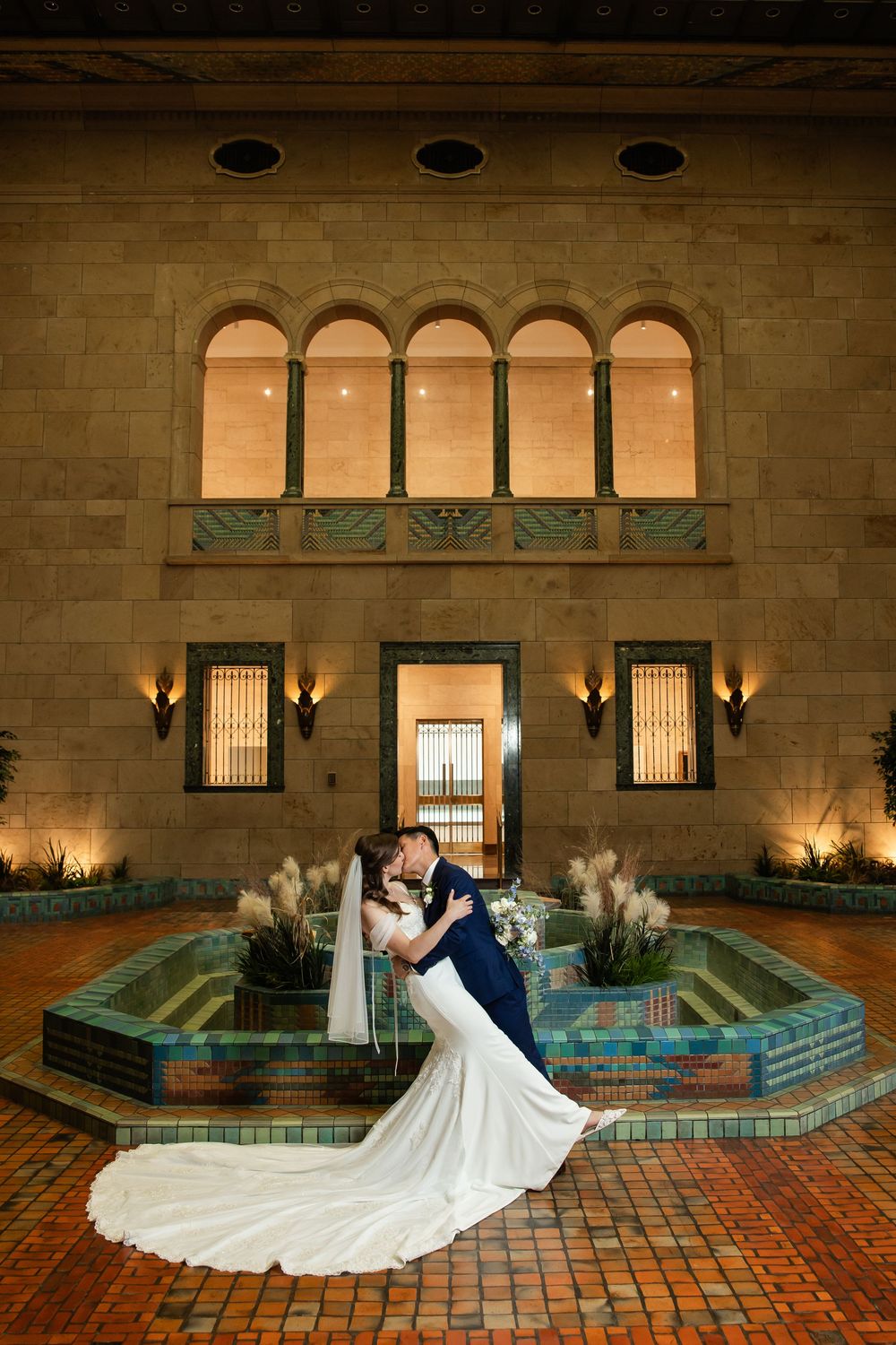 Bride and groom share a romantic dip kiss in front of a fountain at an elegant wedding venue at night.
