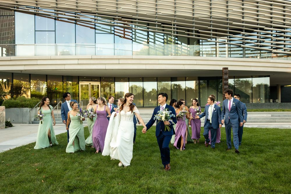 Wedding party walks on green lawn outside modern glass building, bridesmaids in purple and green gowns, groomsmen in blue suits.