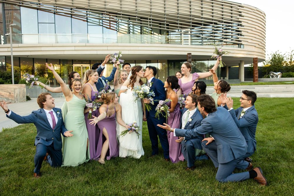 Wedding party celebrating outdoors, bridesmaids in green and purple gowns, groomsmen in navy suits, couple kissing at center.