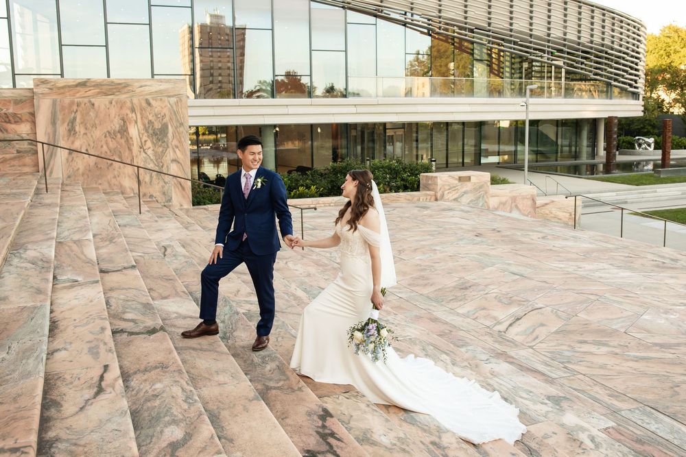 Bride and groom walk hand-in-hand on marble steps outside a modern glass building during golden hour.