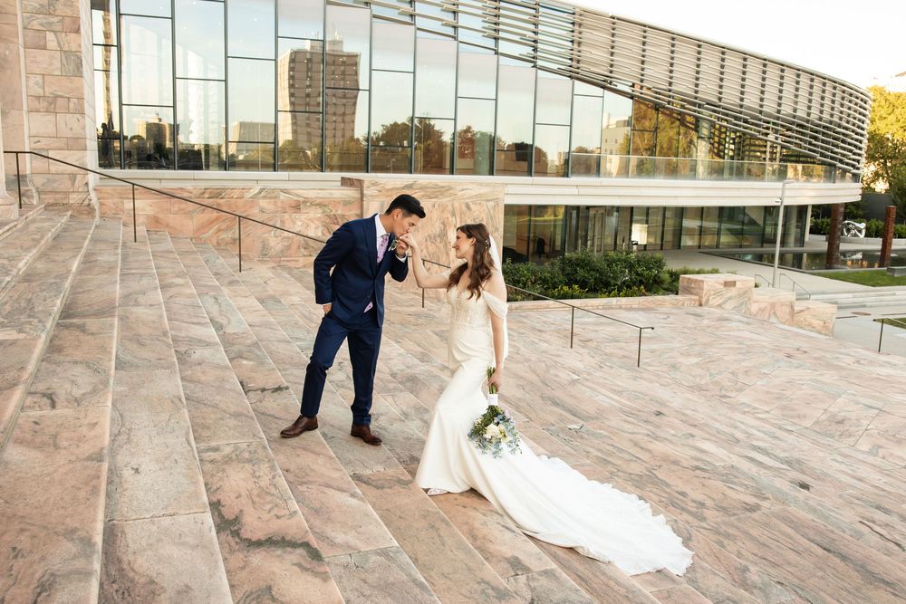 Groom in navy suit kisses bride's hand on stone steps outside modern glass building during golden hour.