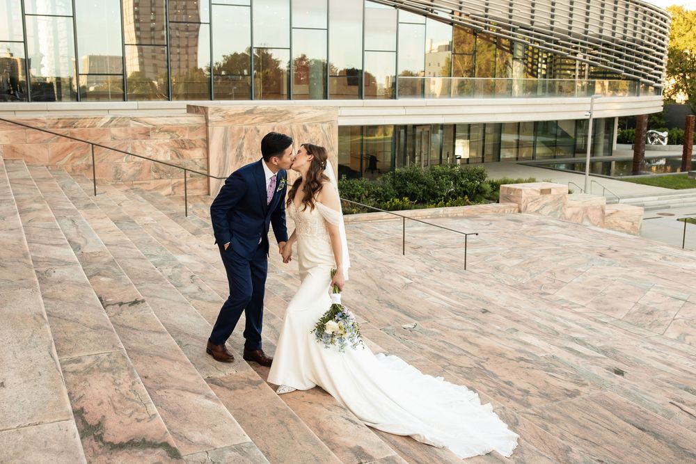 Bride and groom share a romantic kiss on sunlit stone steps with a modern glass building backdrop.