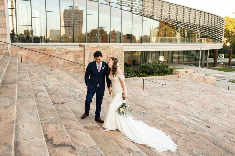 Bride in white gown and groom in navy suit walk hand in hand on stone steps near glass building.