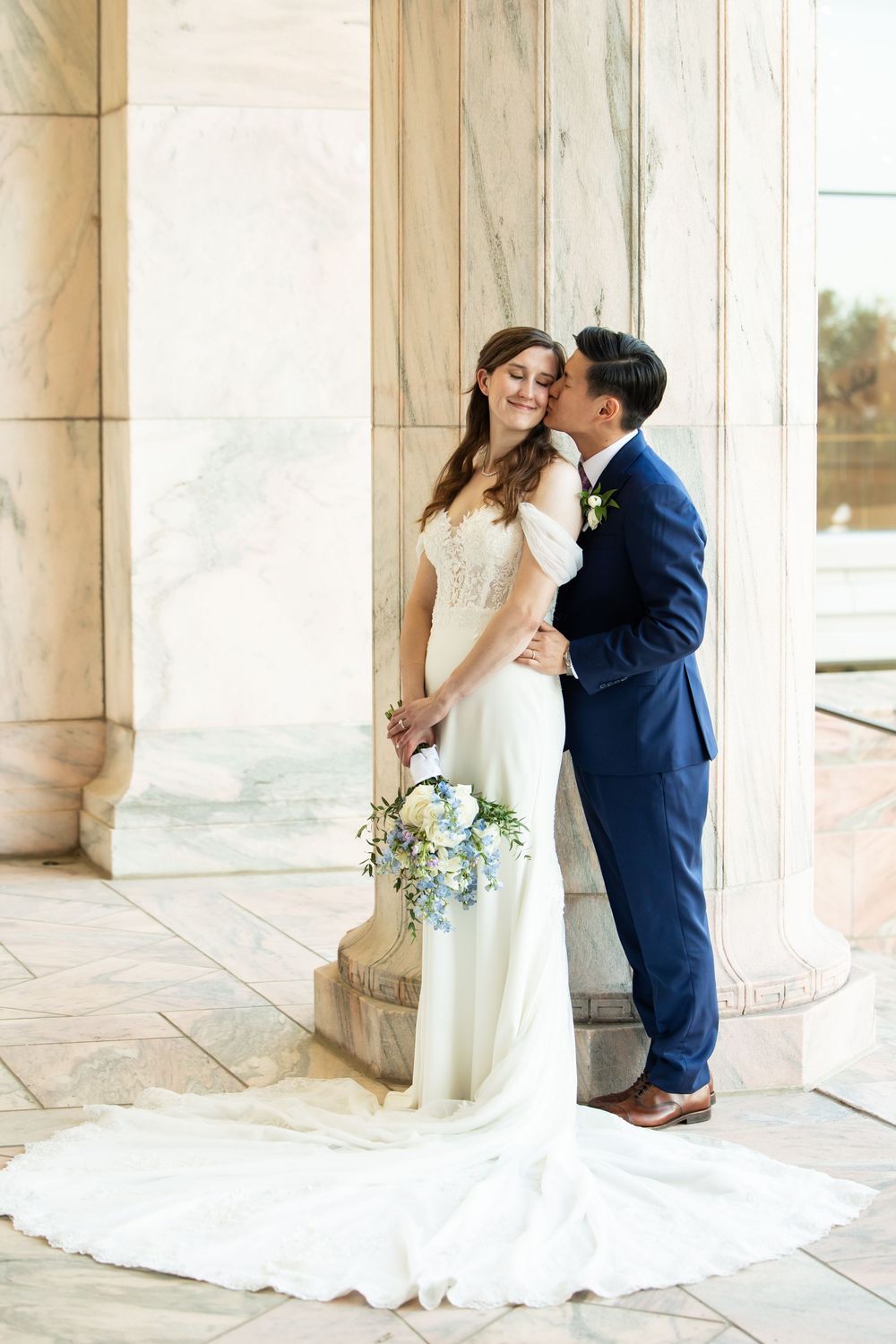 Bride in white gown holds bouquet as groom in navy suit kisses her cheek beside marble columns.