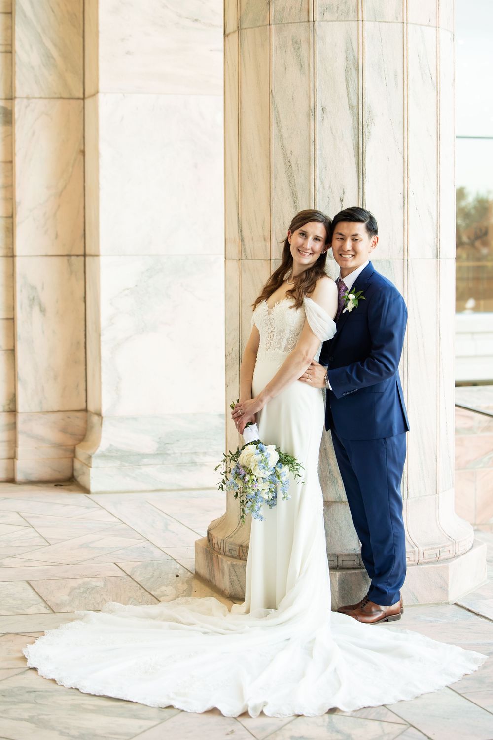 Bride and groom posing between marble columns, she holds a blue and white bouquet, he wears a navy suit.