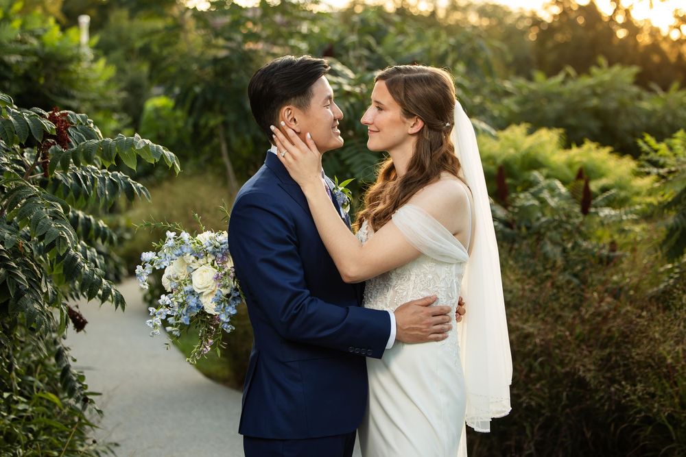 Bride and groom embrace outdoors at golden hour, surrounded by lush greenery, bride holding blue and white bouquet.