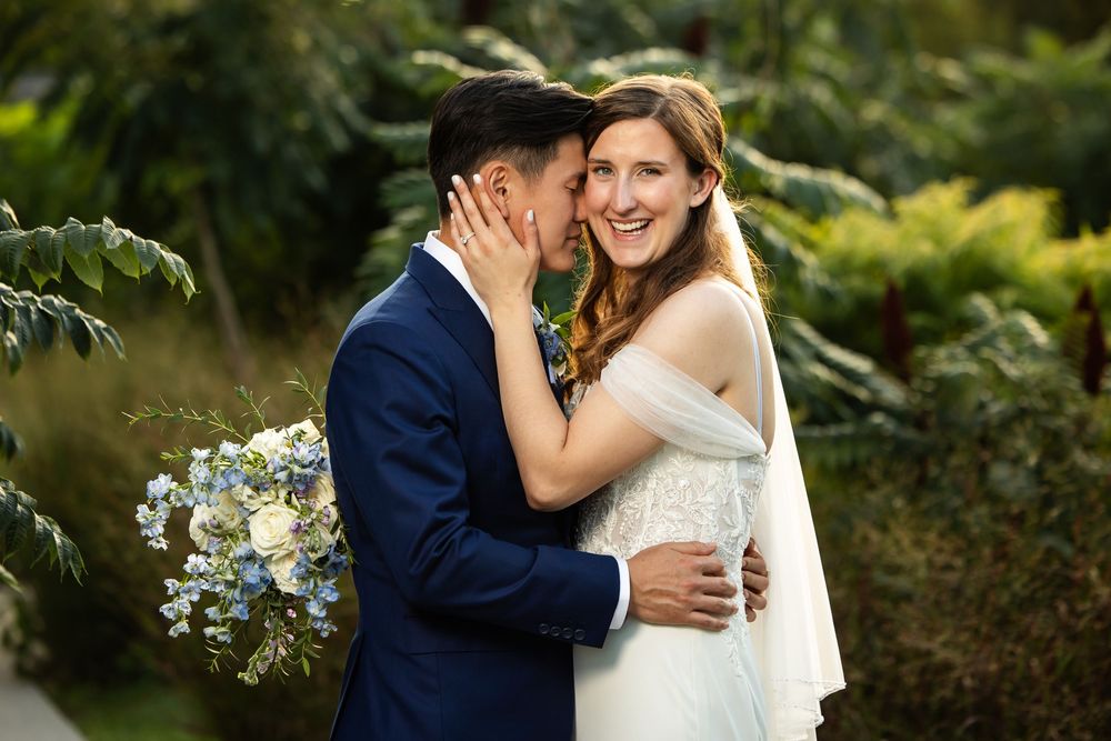 Bride and groom share a tender moment outdoors, with the groom kissing her cheek while she smiles, holding blue flowers.
