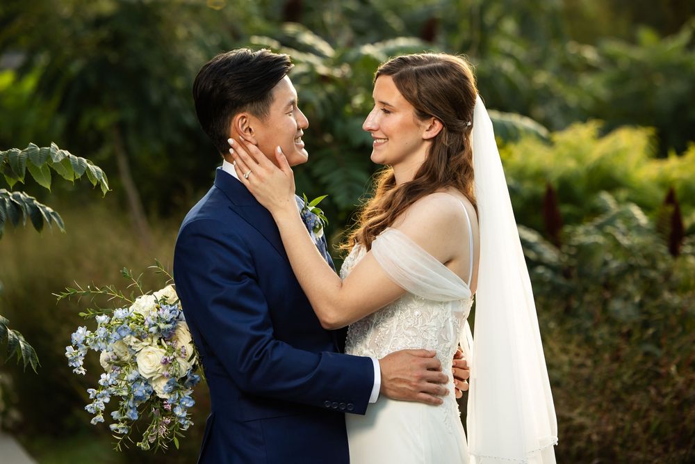 A bride and groom share a tender moment outdoors, with the bride gently holding the groom's face amid lush greenery.