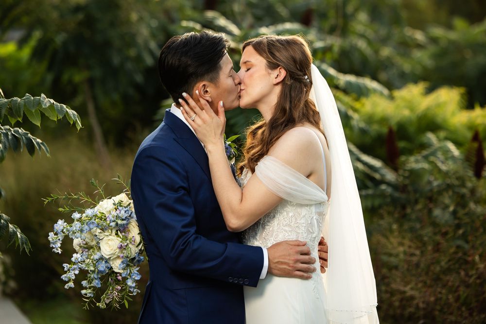 Bride and groom share a romantic kiss outdoors, with bride holding blue floral bouquet, groom in navy suit.