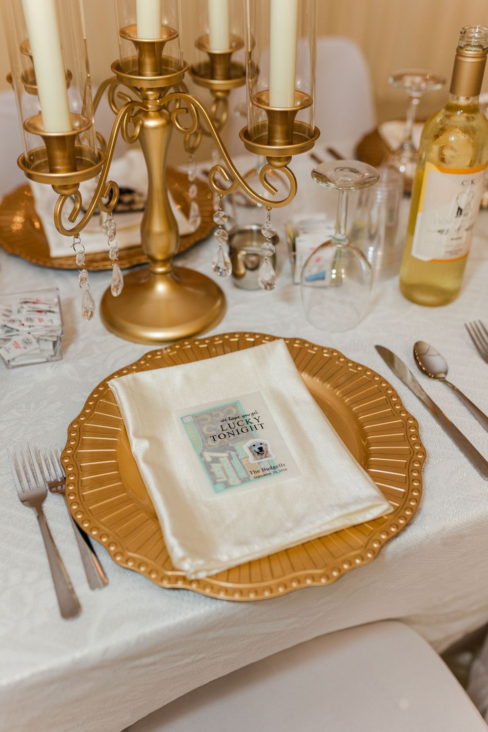 Elegant wedding table setting with gold charger plate, white napkin, candleholder, wine bottle, and crystal glassware on white tablecloth.