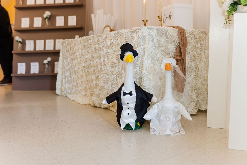 Two decorative geese dressed as bride and groom wedding figurines displayed on a white floor at a wedding venue.