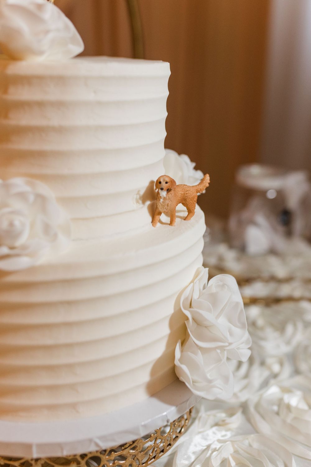 White tiered wedding cake with horizontal ridges, white rose decorations, and a small golden retriever dog figurine topper.