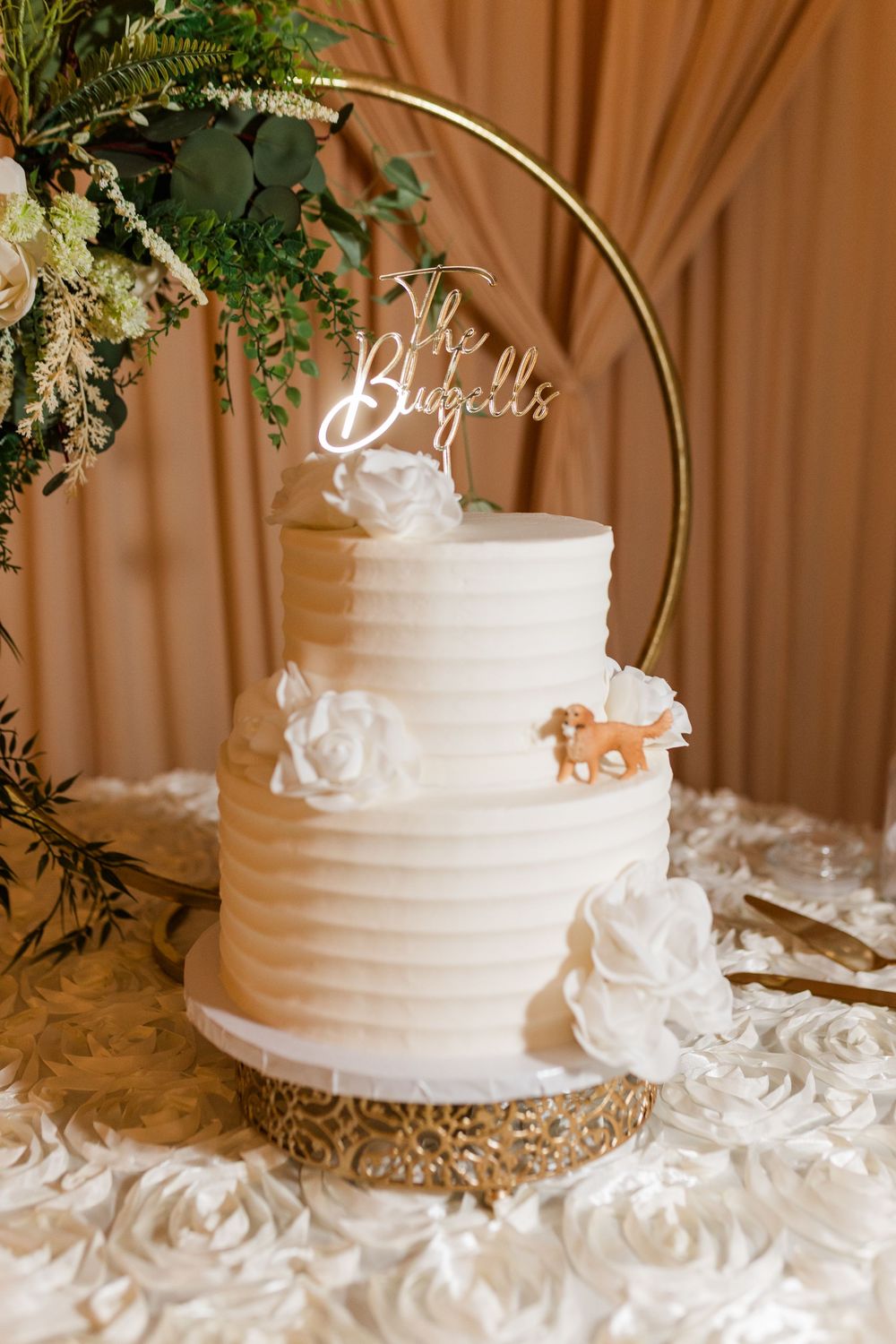 Two-tier white ribbed wedding cake with floral decor, dog figurine, and gold hoop topper reading The Bluzzells.