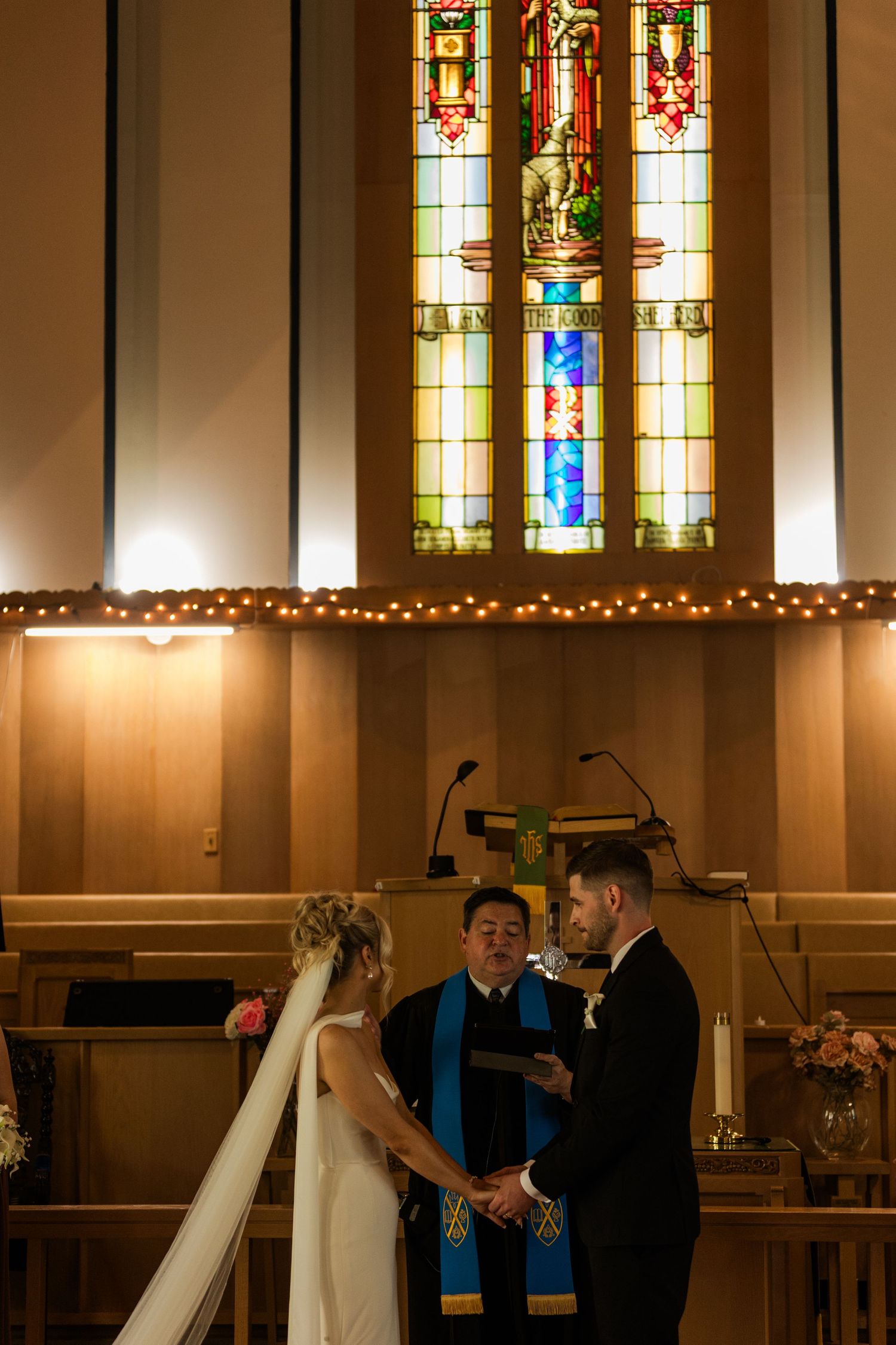 Bride and groom exchange vows at church altar with stained glass windows and officiant during wedding ceremony.