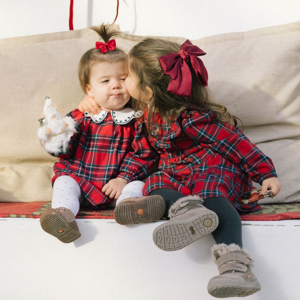 Two little girls in red plaid dresses and bows sit on a cushioned bench as one kisses the other's cheek while holding a small stuffed toy.