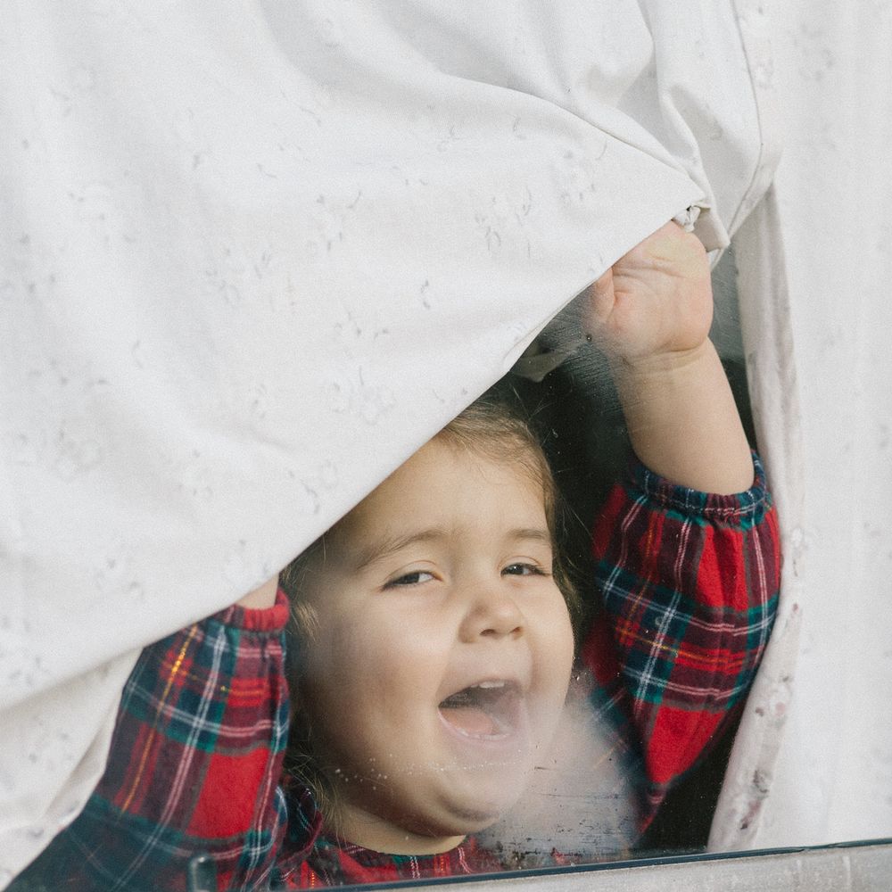 Happy toddler in a red plaid shirt peeks through a window with one hand raised, pulling back a white curtain and smiling.