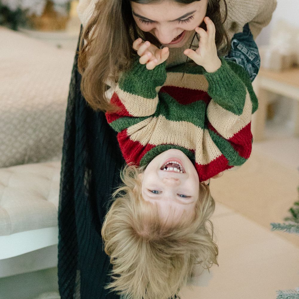 Smiling child in a red, green and cream striped sweater held upside down and laughing while playing with a woman in a bright cozy room.