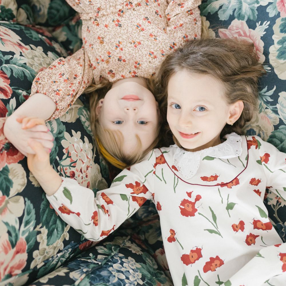Two young girls in floral print dresses lie side by side on a floral cushion, holding hands and smiling up at the camera.