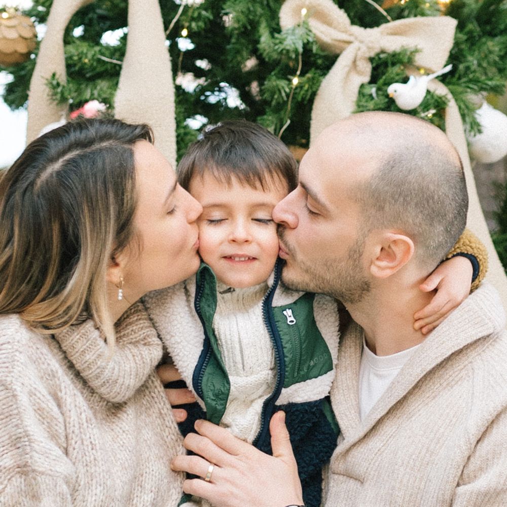 Parents kiss their smiling young son on both cheeks in front of a decorated Christmas tree with burlap ribbon and ornaments.