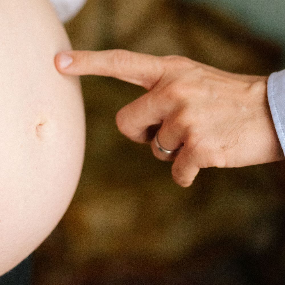 Close-up of a hand with a wedding ring pointing at a pregnant woman's bare belly, showing the belly button.