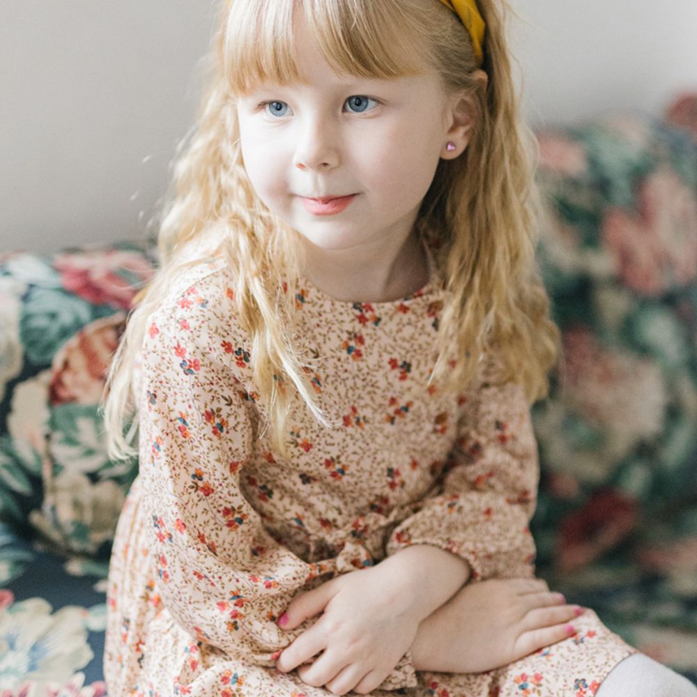 Young girl with long blonde hair and a yellow headband sits on a floral sofa in a ditsy floral dress, hands crossed.
