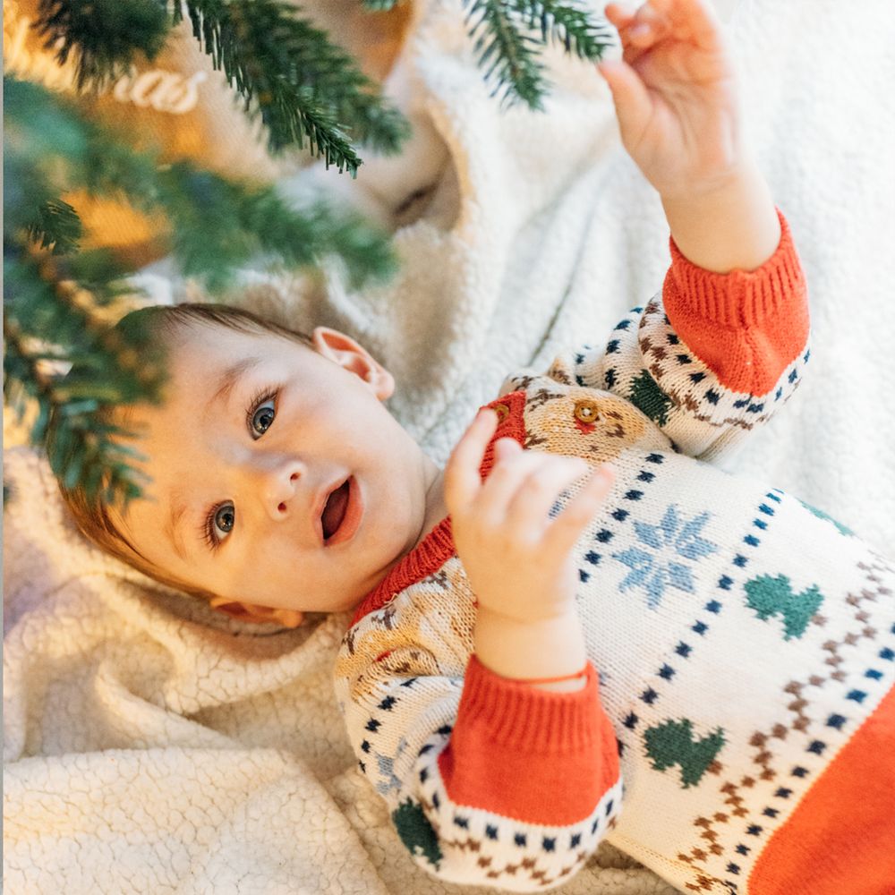 Baby in a festive red and white Christmas sweater with tree patterns lies on a cozy blanket, gazing up at evergreen pine branches.