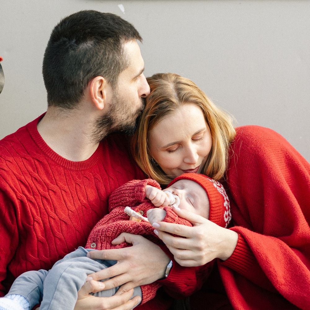 A bearded man kisses the head of a woman in a red sweater as she gently cradles a sleeping newborn baby in a red knit outfit.