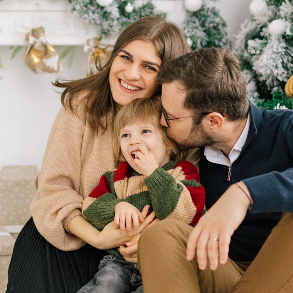 Smiling family of three cuddling by a decorated Christmas tree, with a young child in a striped holiday sweater sitting between mom and dad.