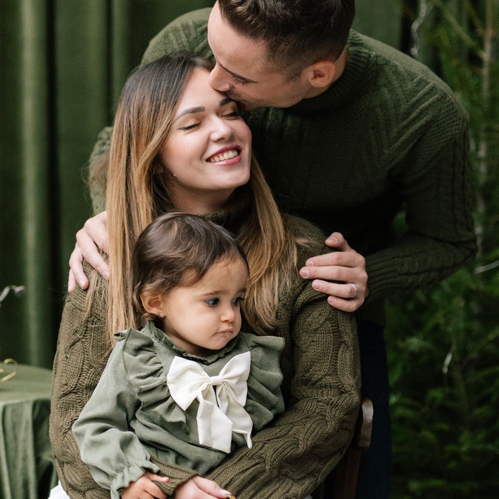 A smiling woman in an olive knit sweater holds a toddler in a ruffled green dress with a white bow while a man kisses her cheek.