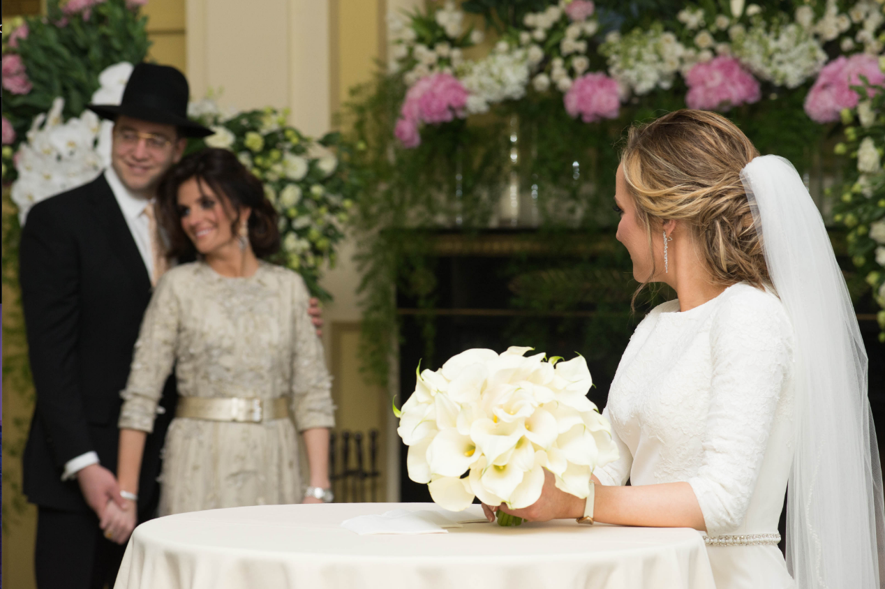 Bride in a white gown and veil holds a bouquet of white calla lilies at a table as a couple smiles behind her in a floral-decorated venue.