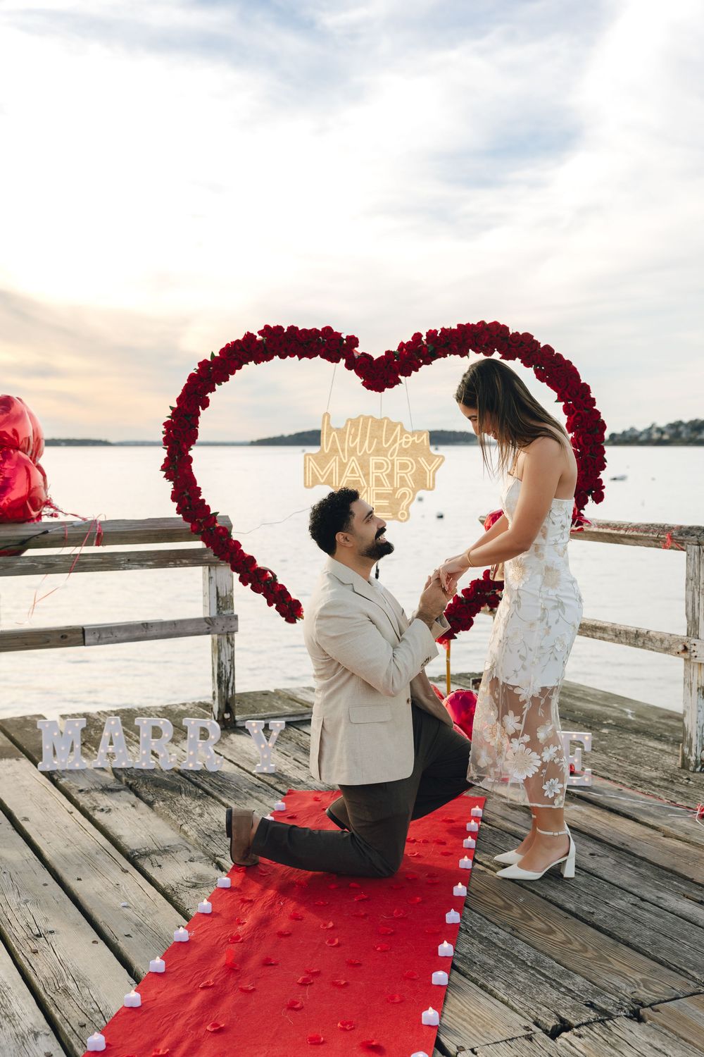 Man proposes to woman on one knee beside a red rose heart arch with Will You Marry Me sign on a dock.