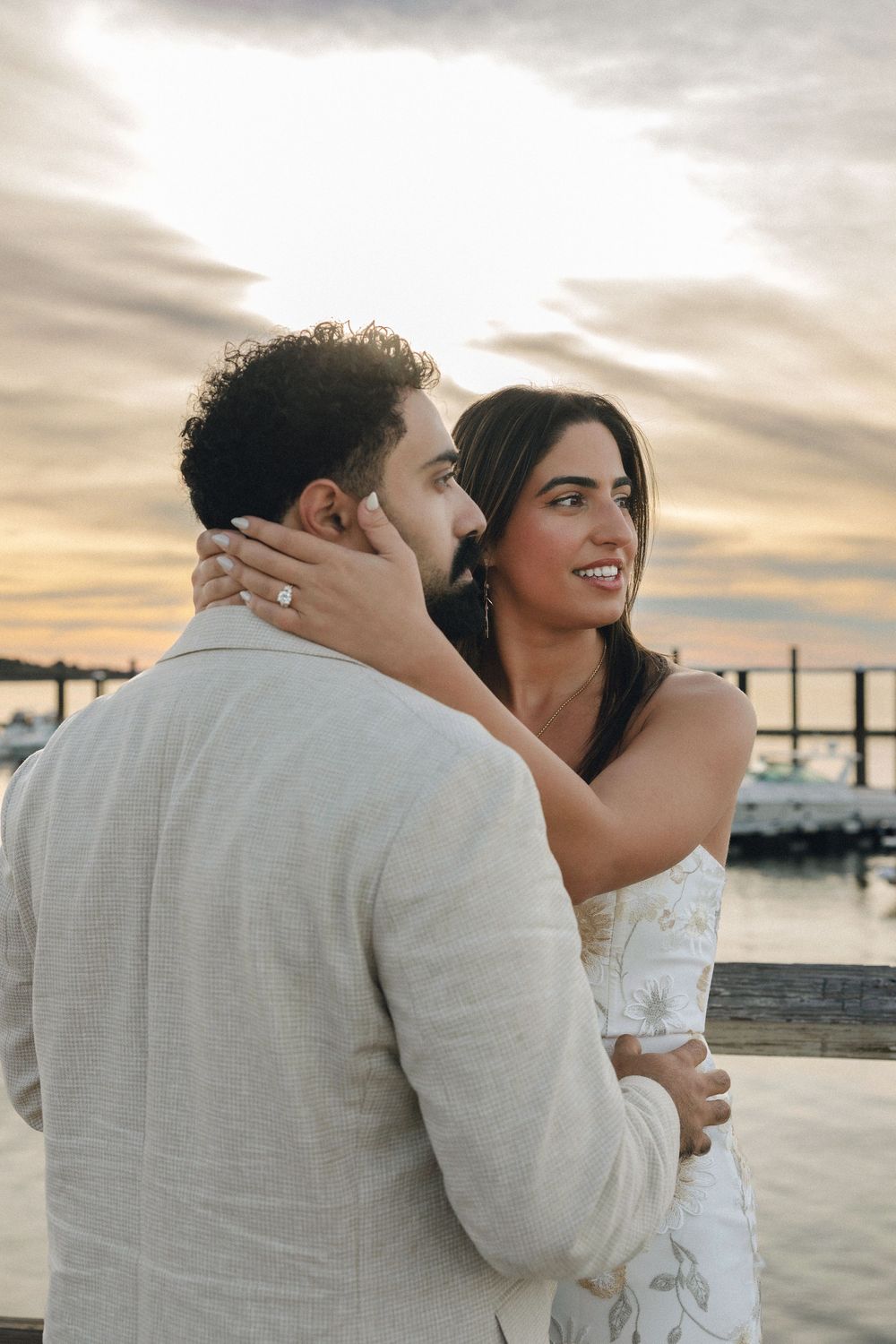 Couple embracing at sunset near a marina, woman in white dress touching man's face in a romantic outdoor portrait.
