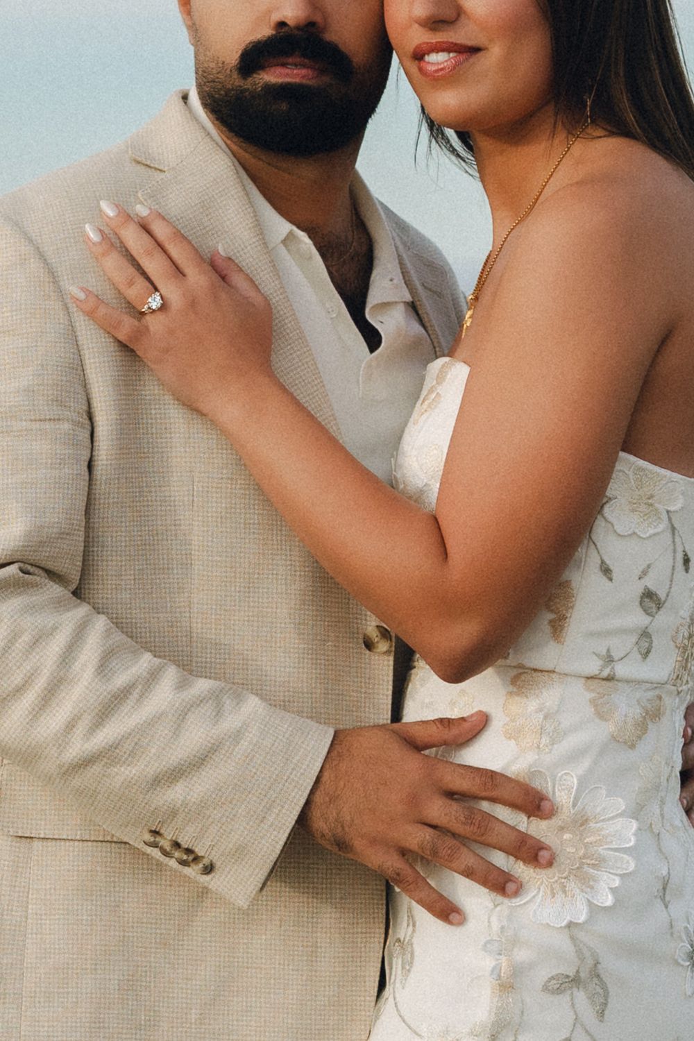 Couple in wedding attire embracing, bride in floral dress with diamond ring, groom in beige suit.